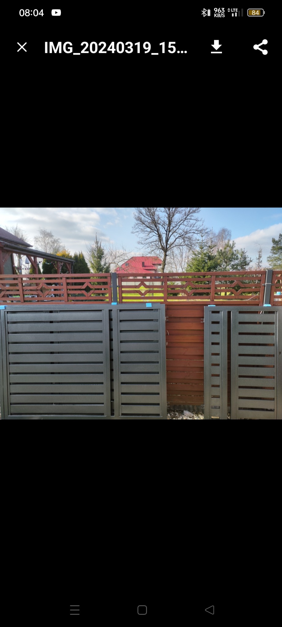 Szare, poziome panele ogrodzeniowe, częściowo zmontowane, z widocznym tłem z drewnianych desek i brązowego ogrodzenia z geometrycznym wzorem, ustawione na żwirowym podłożu.