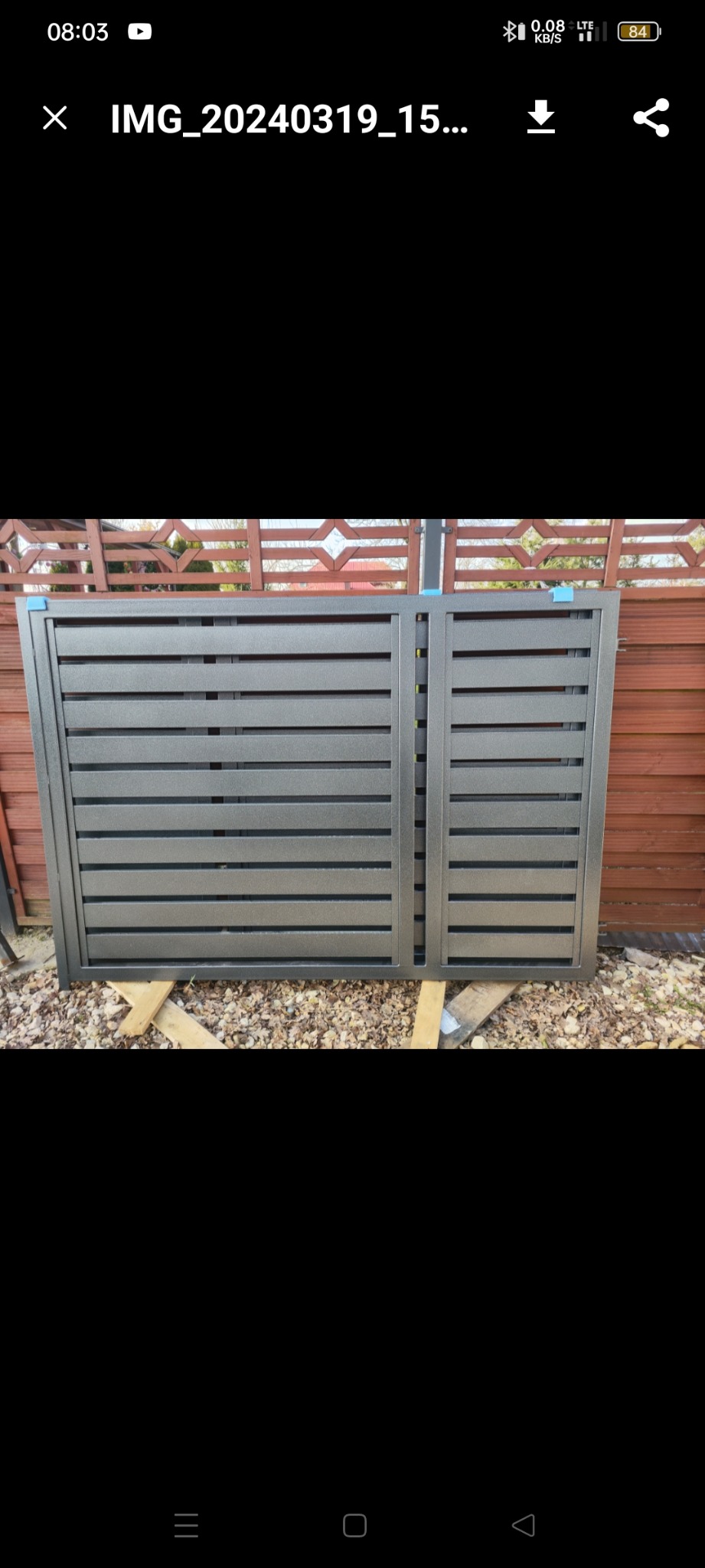 Fragment szarej, metalowej bramy wjazdowej z poziomymi panelami, oparty na drewnianych belkach na zewnątrz, z widocznym drewnianym ogrodzeniem w tle.