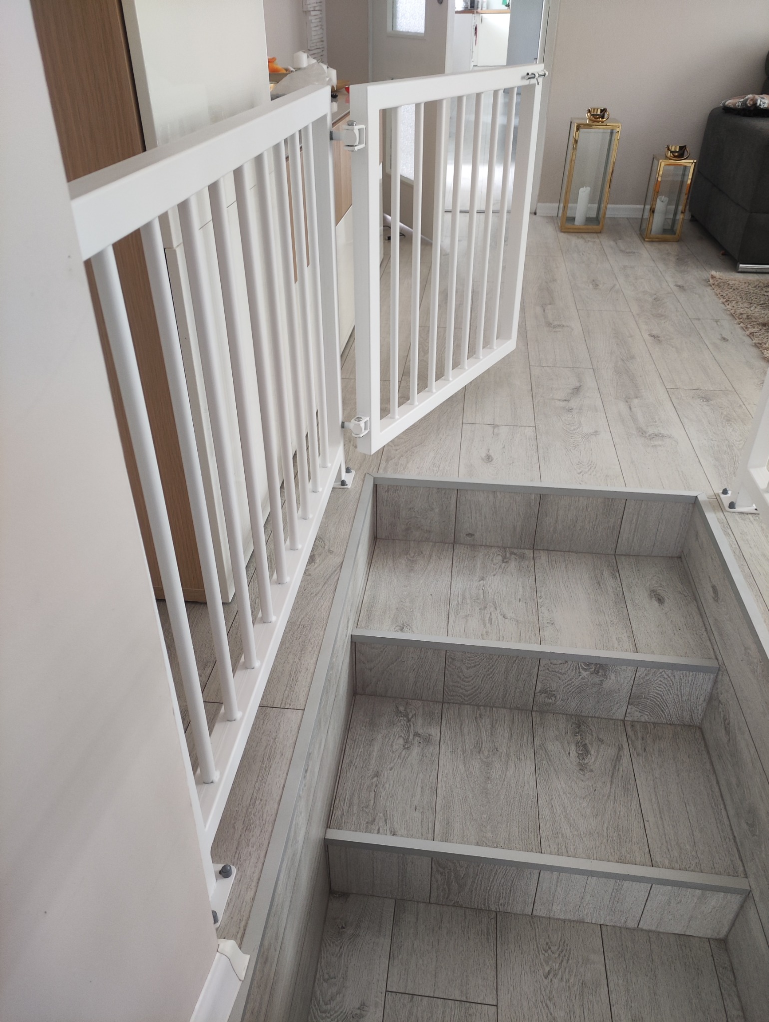 Biała barierka zabezpieczająca schody wewnętrzne z drewnopodobnymi stopniami, widoczna z perspektywy góry, z częściowo otwartymi drzwiczkami. W tle elementy wystroju wnętrza.