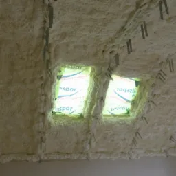 Poddasze z dwoma oknami dachowymi zabezpieczonymi folią, pokryte grubą warstwą jasnej pianki izolacyjnej, z widocznymi metalowymi elementami mocującymi.