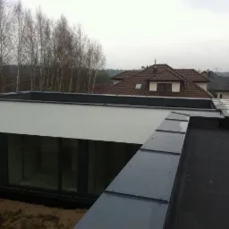 Nowoczesny dach płaski z obróbką blacharską w kolorze grafitowym. W tle widoczny budynek z dachem skośnym pokrytym brązową dachówką ceramiczną.