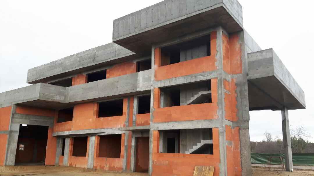 Surowy stan dwupiętrowego budynku z pustaków ceramicznych i betonu, widoczne schody wewnętrzne, teren budowy w tle.