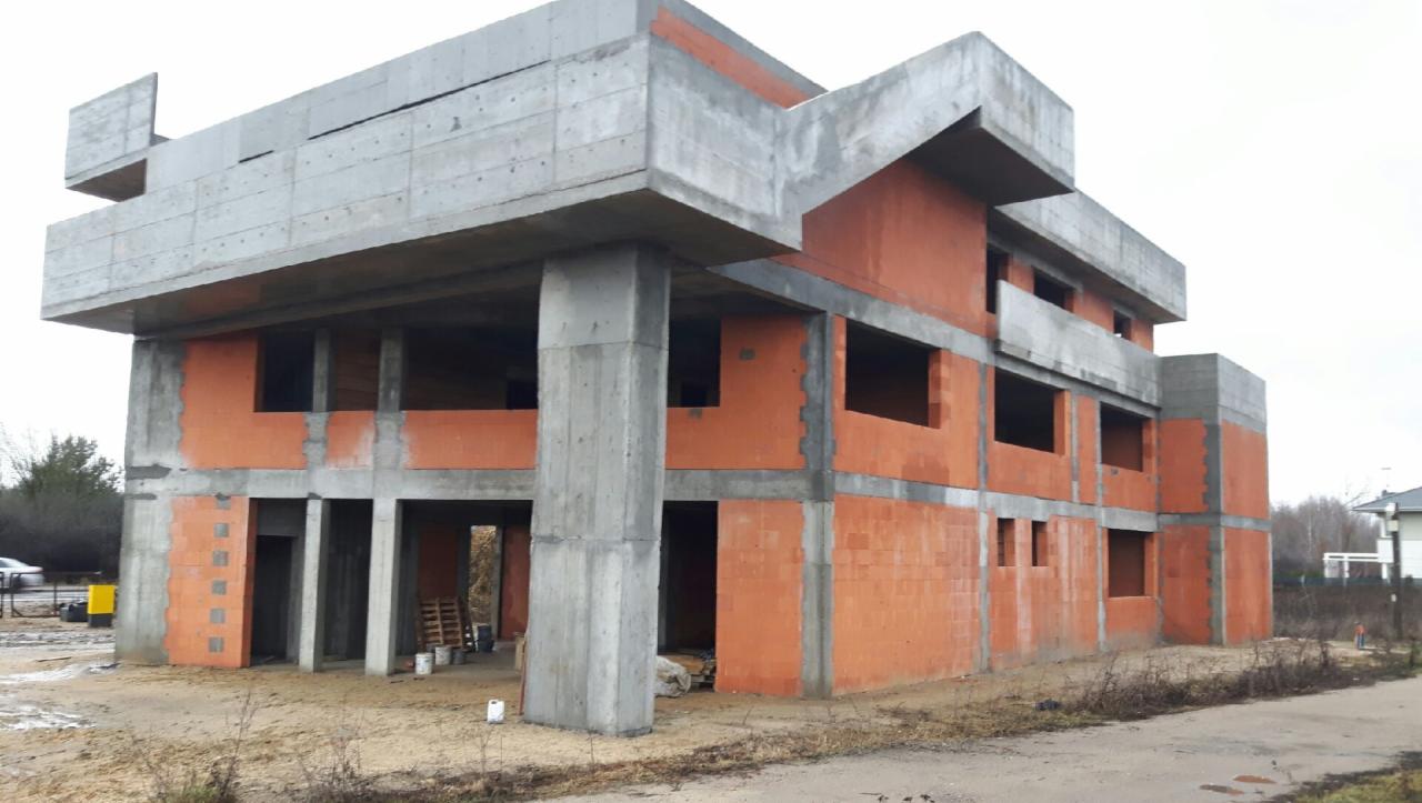Surowy stan dwupiętrowego budynku w trakcie budowy z betonu i cegły ceramicznej, widoczne niezadaszone otwory okienne i drzwiowe, zarys konstrukcji dachu.
