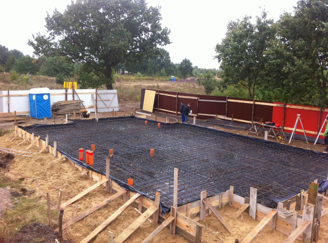 Zbrojenie fundamentu domu jednorodzinnego z widoczną szalunką, izolacją i częściowo ogrodzeniem. W tle drzewa i przenośna toaleta.