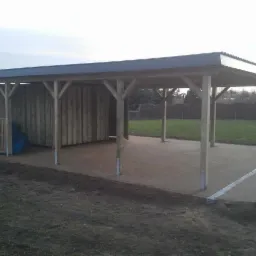 Drewniana konstrukcja z zadaszeniem, częściowo zabudowana, z podłożem z kostki brukowej, widoczne worki w części zabudowanej.