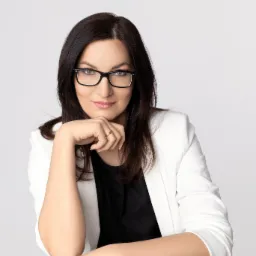 Portret kobiety w okularach, białej marynarce i czarnej bluzce, opierającej podbródek na dłoni, z delikatnym uśmiechem na jasnym tle.