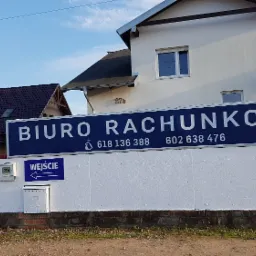 Dwupiętrowy budynek z dużym niebieskim szyldem z napisem 'BIURO RACHUNKOWE' i numerami telefonów, mały szyld 'WEJŚCIE' z białą strzałką, skrzynka pocztowa i dom w tle z ceglanym kominem.