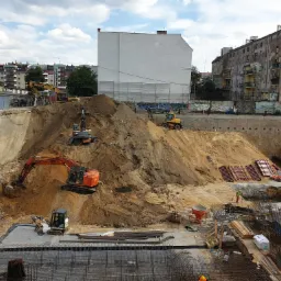 Rozległy plac budowy z kilkoma pracującymi koparkami i robotnikami, widok z góry na wykop z widocznym zbrojeniem i elementami konstrukcyjnymi.