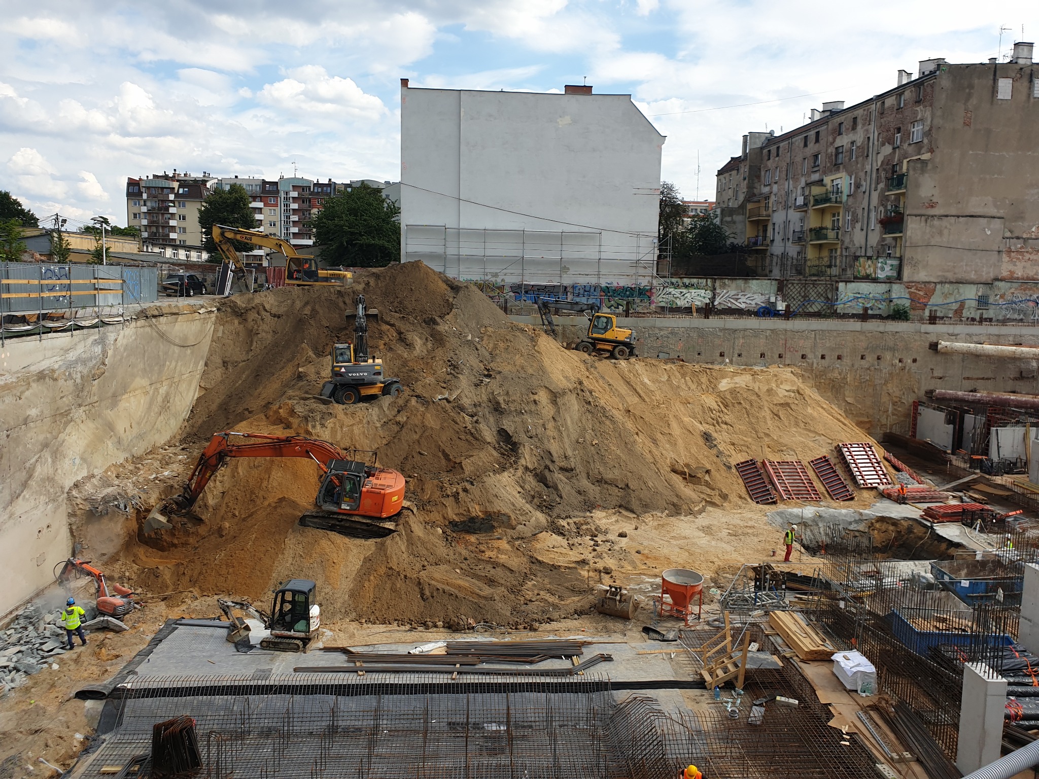 Rozległy plac budowy z kilkoma pracującymi koparkami i robotnikami, widok z góry na wykop z widocznym zbrojeniem i elementami konstrukcyjnymi.