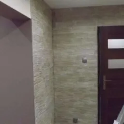 Ściana wykończona dekoracyjnym tynkiem strukturalnym w odcieniach beżu i szarości, z widocznym fragmentem ściany przygotowanej pod malowanie i drabiną opartą o futrynę drzwi.