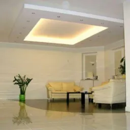 Jasne wnętrze z podwieszanym sufitem z oświetleniem punktowym i centralnym panelem świetlnym, kremowe meble wypoczynkowe, błyszcząca podłoga z płytek, roślina doniczkowa w zielonej osłonce.