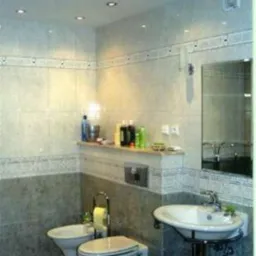 Wykończona łazienka w szaro-białej kolorystyce z podwieszaną toaletą i bidetem, umywalką na ozdobnym stelażu i półką z kosmetykami, oświetlona punktowymi lampami w suficie.