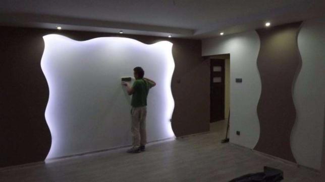 Pracownik wygładza ścianę z podświetlanym, falistym wykończeniem w kolorze białym, sąsiadującym z brązowymi, dekoracyjnymi elementami na ścianie. Widoczne jest także oświetlenie sufitowe punktowe.