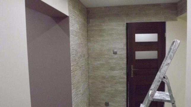 Ściana wykończona dekoracyjnym tynkiem strukturalnym w odcieniach beżu i szarości, z widocznym fragmentem ściany przygotowanej pod malowanie i drabiną opartą o futrynę drzwi.