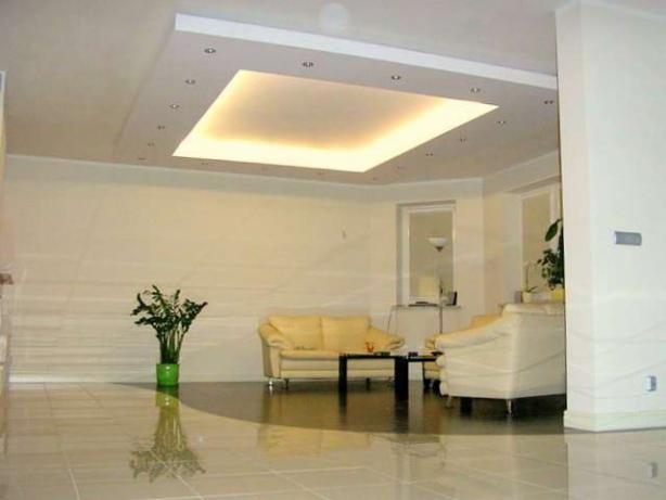 Jasne wnętrze z podwieszanym sufitem z oświetleniem punktowym i centralnym panelem świetlnym, kremowe meble wypoczynkowe, błyszcząca podłoga z płytek, roślina doniczkowa w zielonej osłonce.