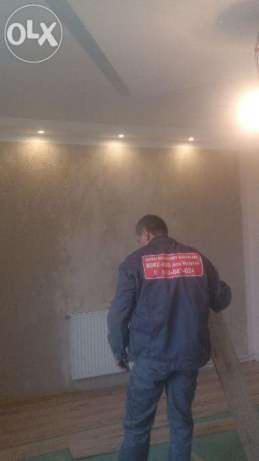 Pracownik w uniformie z logo firmy stoi w pokoju z świeżo położonymi panelami podłogowymi i oświetleniem punktowym na ścianie o nierównej fakturze.