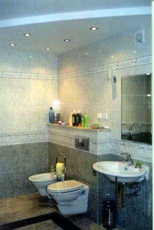 Wykończona łazienka w szaro-białej kolorystyce z podwieszaną toaletą i bidetem, umywalką na ozdobnym stelażu i półką z kosmetykami, oświetlona punktowymi lampami w suficie.