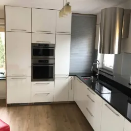 Nowoczesna kuchnia z białymi, lakierowanymi frontami, czarnym blatem, wbudowanym piekarnikiem i okapem ze stali nierdzewnej, oświetlona wiszącą lampą.