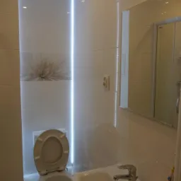 Nowoczesna łazienka z białymi płytkami, podświetleniem LED w pionowej wnęce, lustrem i umywalką. Widoczny fragment toalety. Minimalistyczny design.