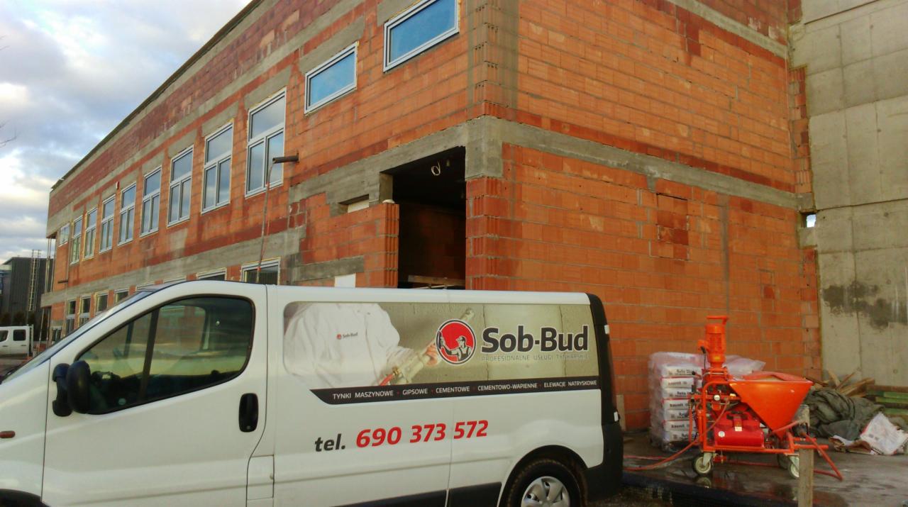 Budynek w trakcie budowy z pustaków, z widocznym autem firmy Sob-Bud i maszyną do tynkowania. Na aucie numer telefonu kontaktowy.