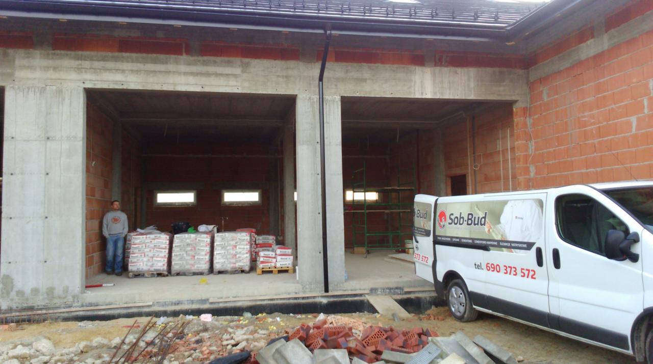 Budynek w stanie surowym otwartym przed tynkowaniem maszynowym, widoczne worki z zaprawą, pracownik i firmowy bus Sob-Bud. Cegła, beton, okna.