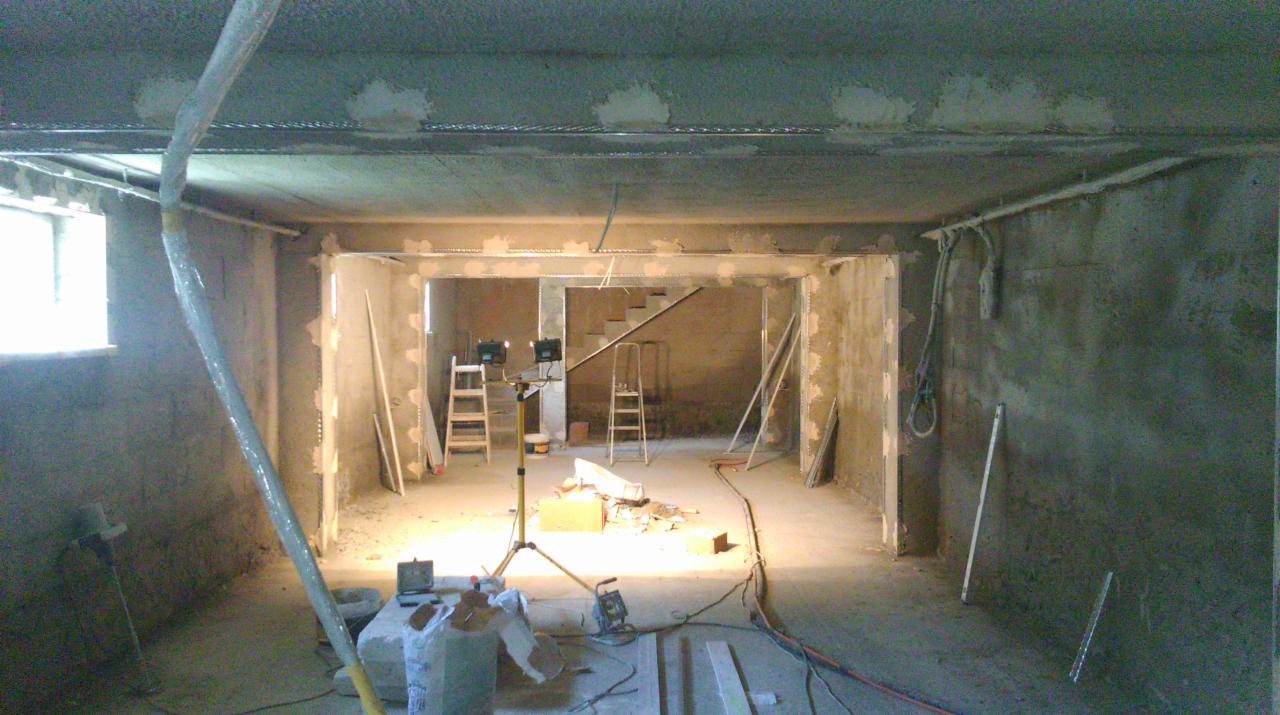 Wnętrze w trakcie prac przygotowawczych do tynkowania: ściany surowe, widoczne narzędzia, drabiny, oświetlenie budowlane, rury instalacyjne na ścianie.