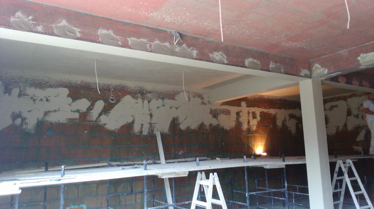 Ściana z cegły częściowo pokryta tynkiem, widoczne rusztowanie i elementy konstrukcyjne budynku w trakcie prac wykończeniowych. Fragment osoby w tle.