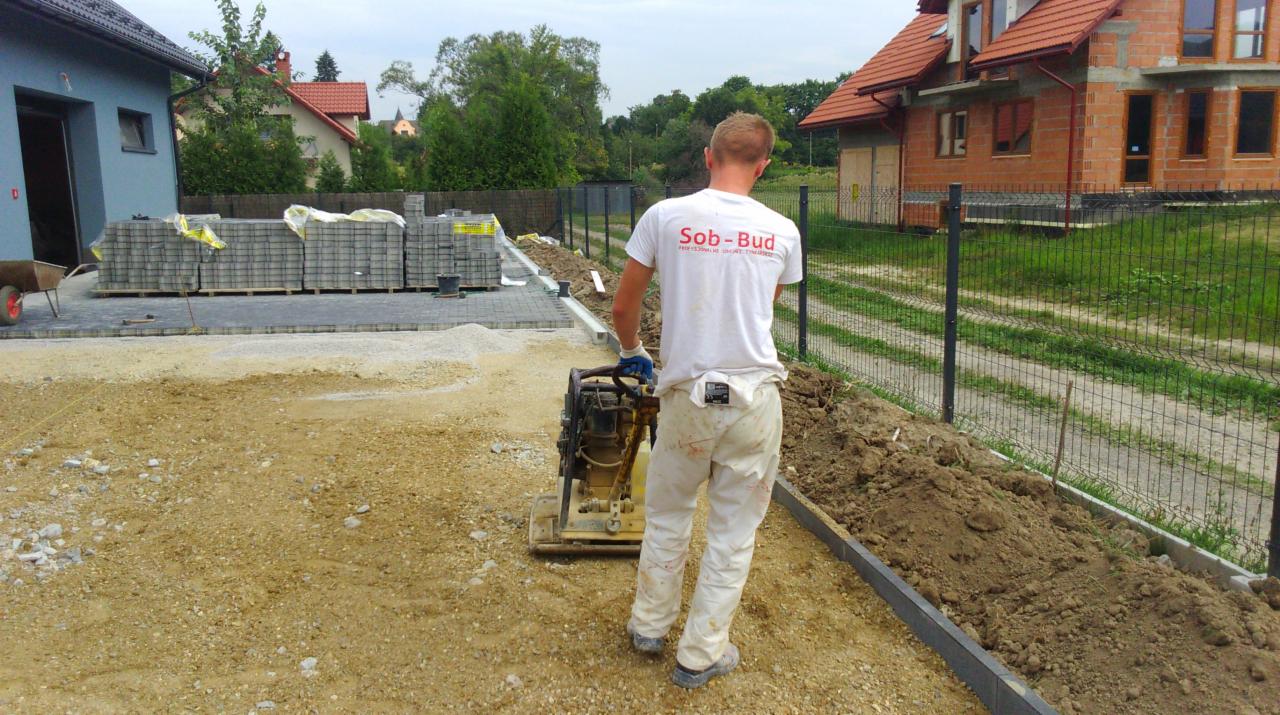 Pracownik ubija ziemię zagęszczarką w przygotowaniu podłoża pod kostkę brukową. W tle ułożona kostka i dom w trakcie budowy.