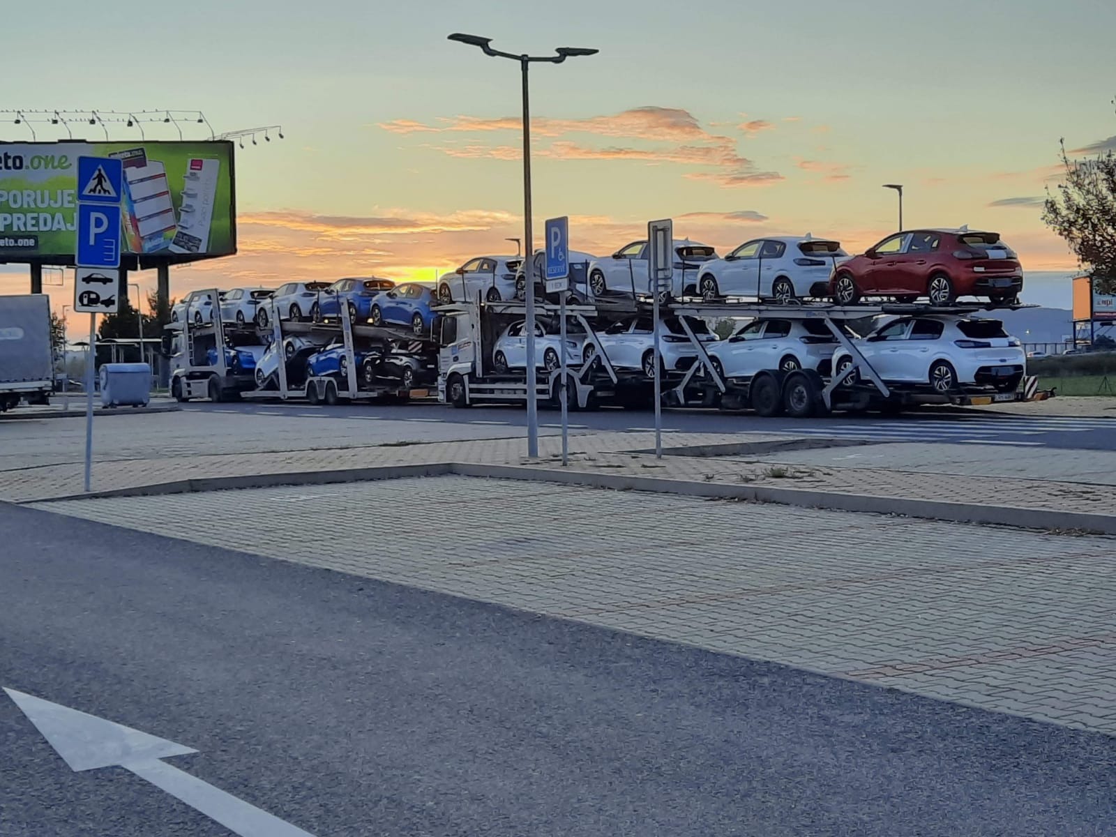 Dwu-poziomowa laweta z załadowanymi samochodami osobowymi, głównie białymi, z jednym czerwonym na najwyższym poziomie, na tle zachodzącego słońca i billboardu.