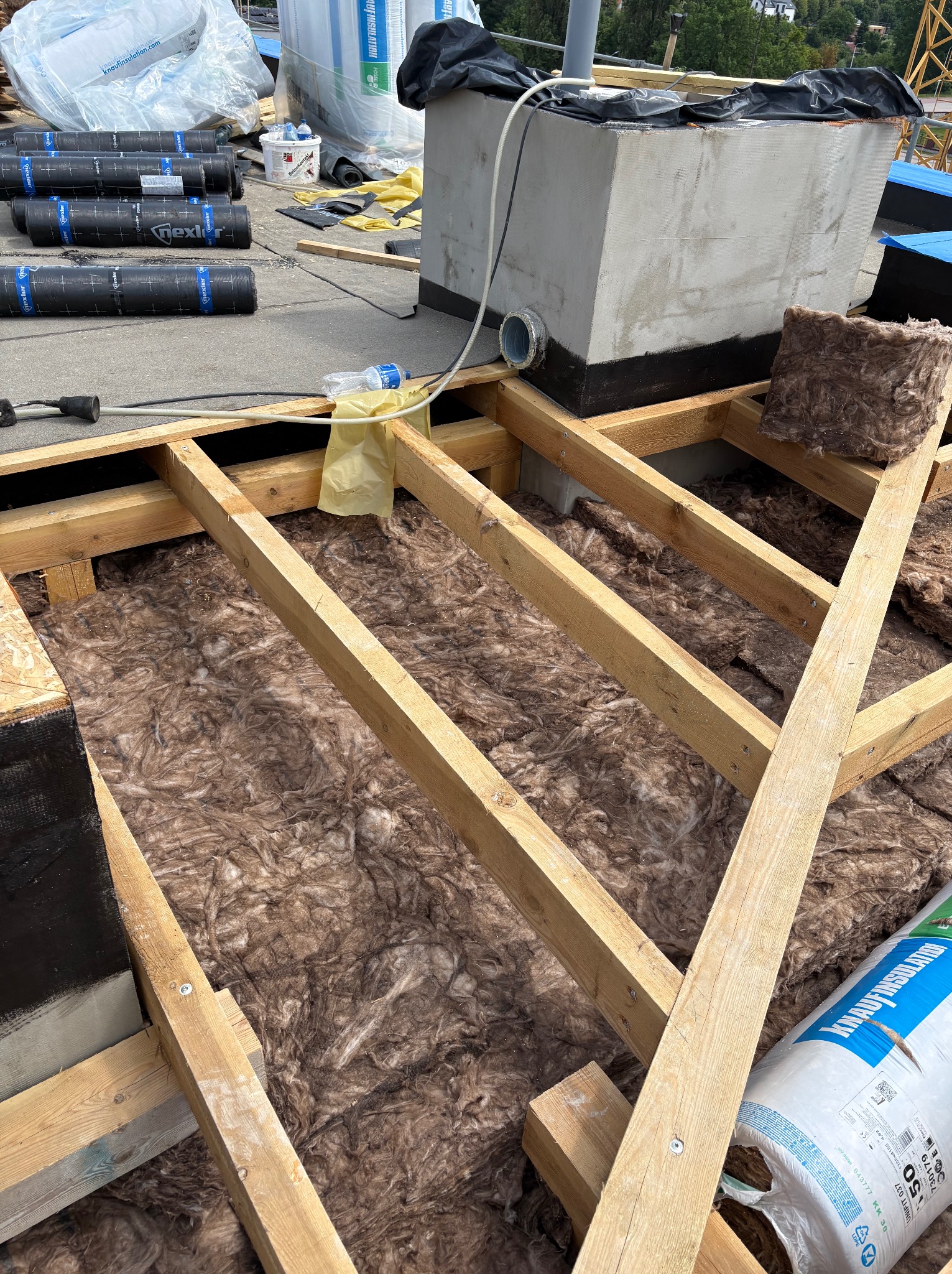 Ocieplenie dachu wełną mineralną, widoczne elementy konstrukcji drewnianej, rolki papy i komin wentylacyjny. Prace dekarskie w toku, izolacja termiczna.