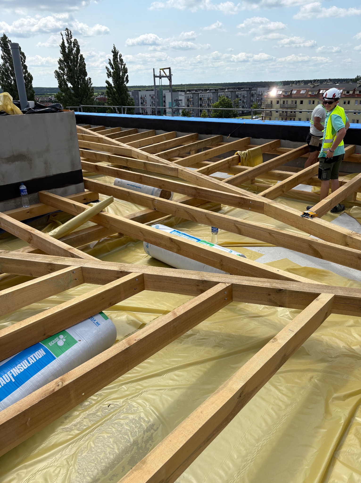 Konstrukcja drewniana dachu pokryta żółtą membraną, widoczne rolki izolacji i pracownicy w oddali. Dach w trakcie budowy, widok z góry, słoneczny dzień.