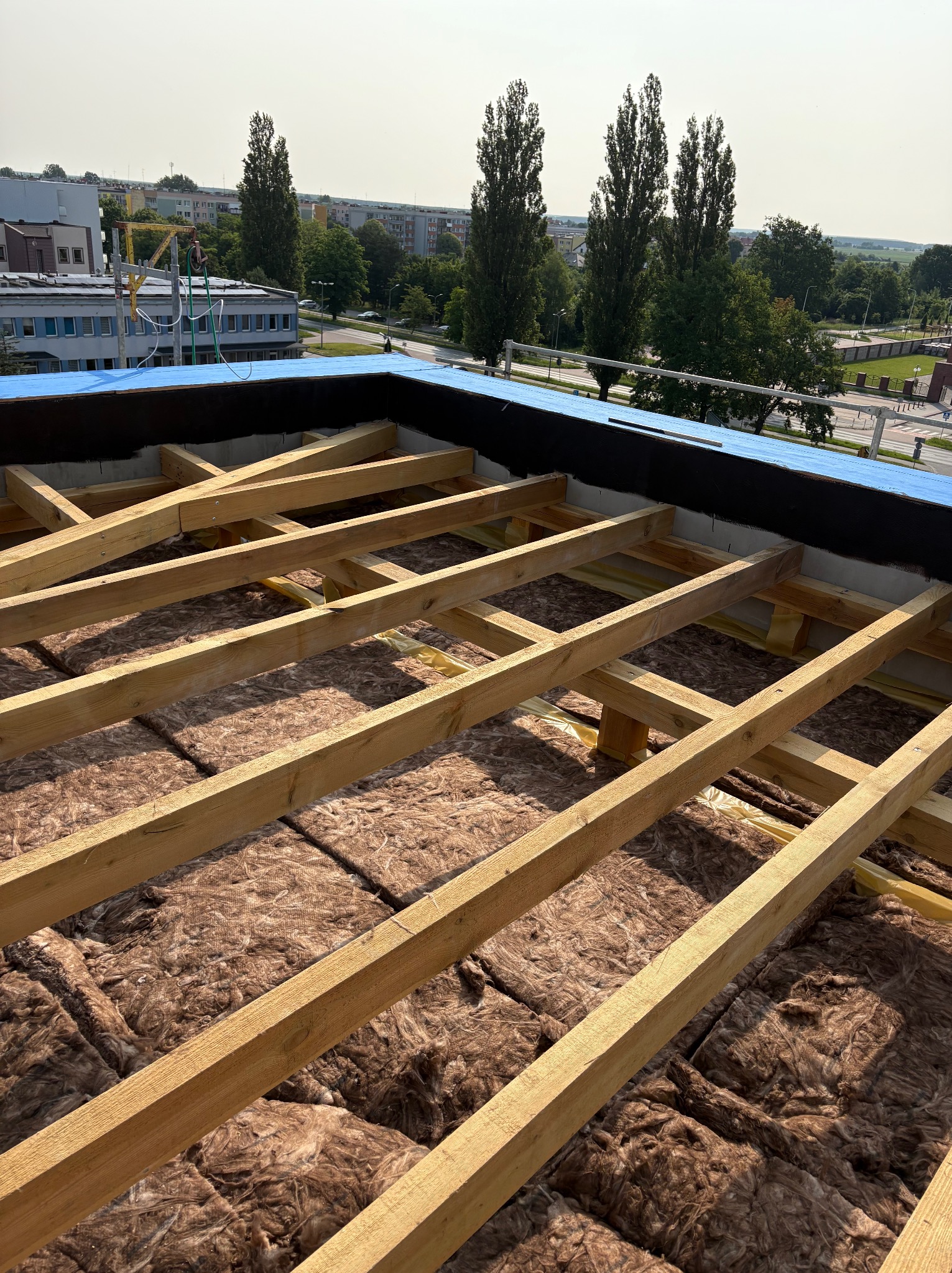 Montaż izolacji termicznej z wełny mineralnej i drewnianej konstrukcji więźby na dachu płaskim budynku. Widok z góry, w tle zabudowa miejska i drzewa.