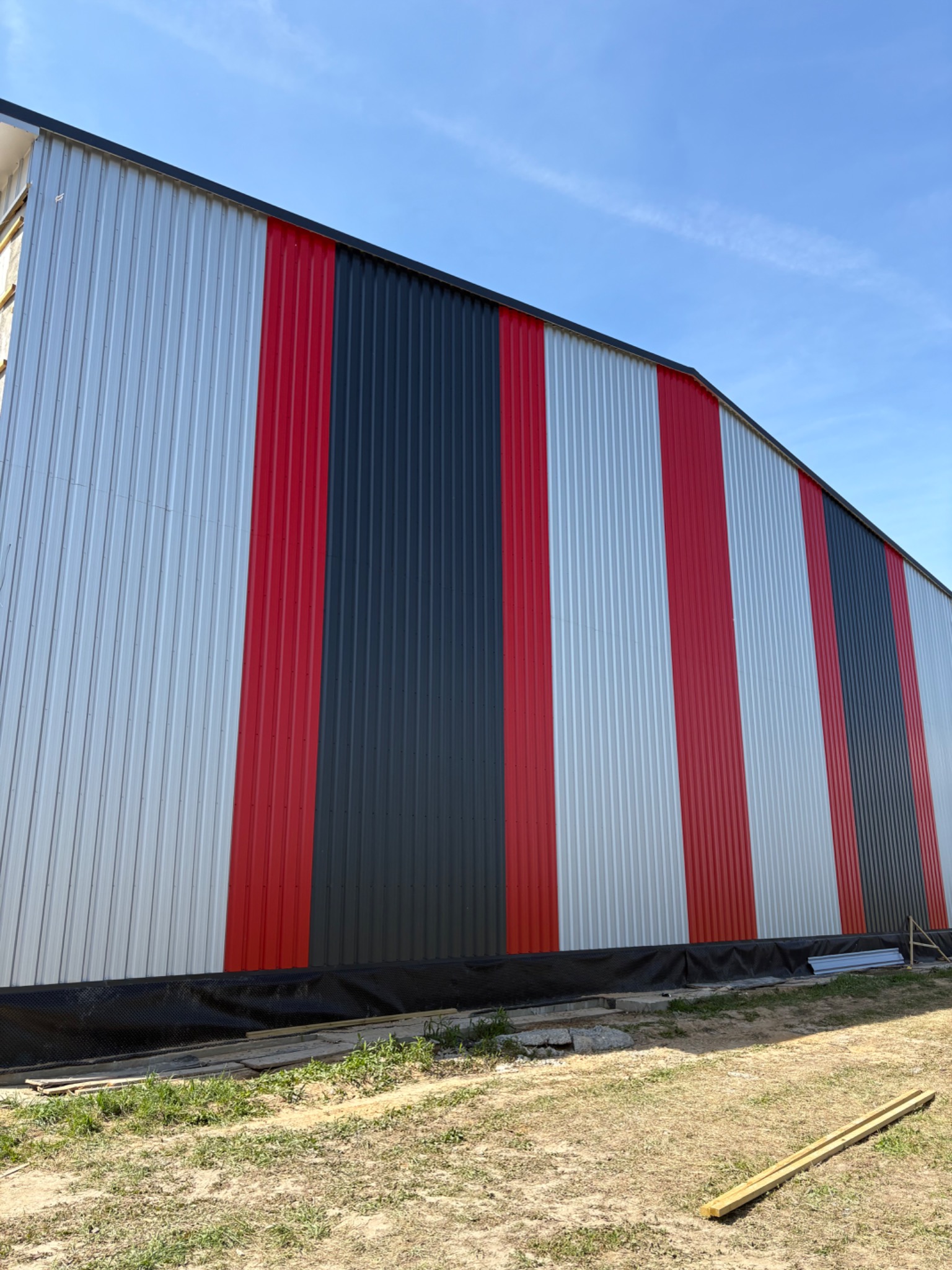 Kolorowa elewacja hali z blachy trapezowej w pasy: srebrne, czerwone i grafitowe. Widok z dołu, błękitne niebo w tle. Nowoczesne wykończenie budynku.