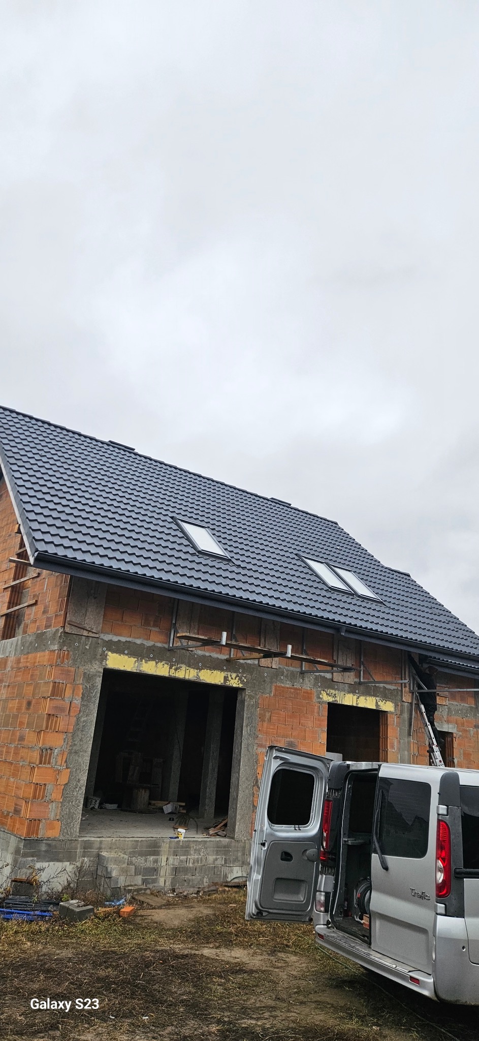 Dom w trakcie budowy z zamontowanym, grafitowym dachem z trzema oknami dachowymi. Na pierwszym planie samochód dostawczy z otwartymi drzwiami.