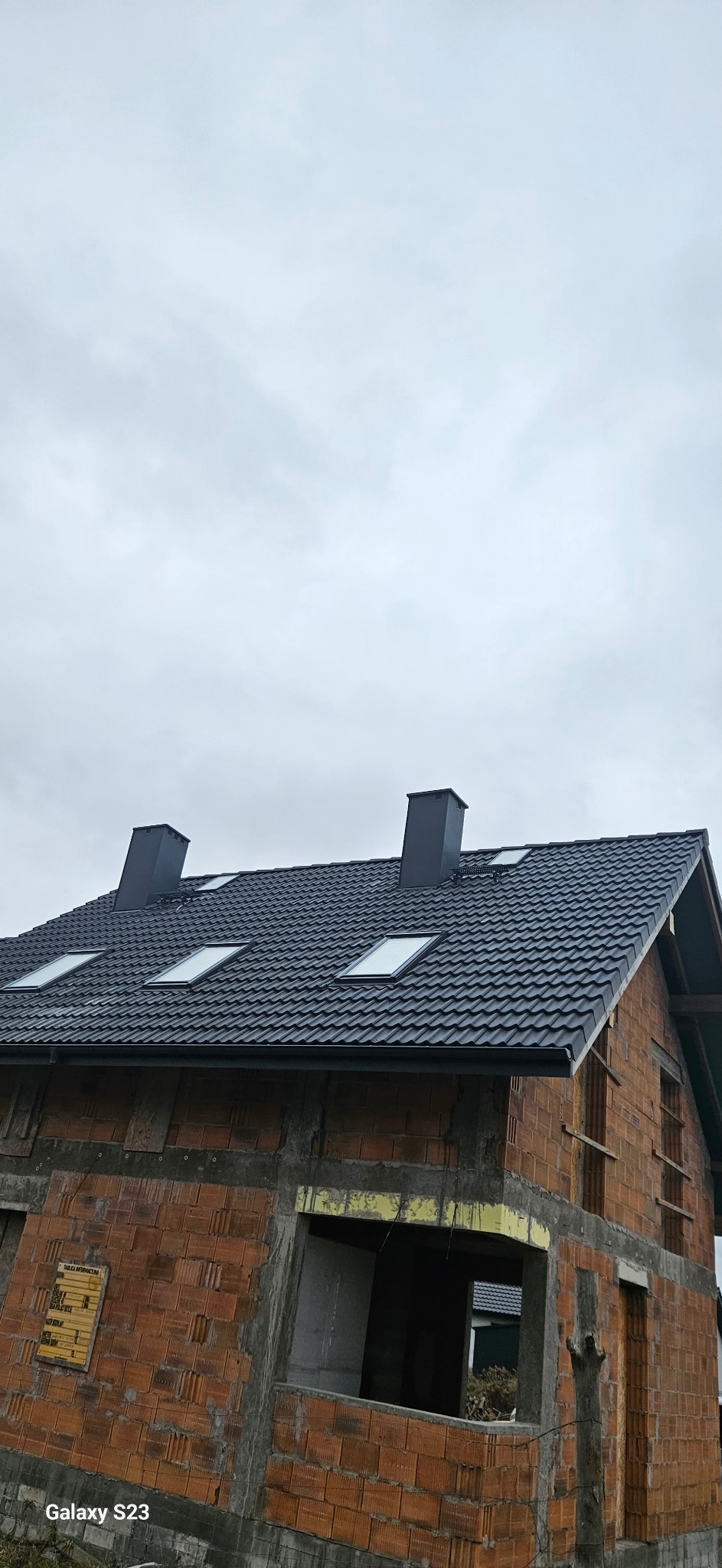 Ciemna dachówka ceramiczna na nowym domu z oknami dachowymi i kominami. Budynek w stanie surowym otwartym, z widoczną konstrukcją z cegły. Pochmurne niebo w tle.