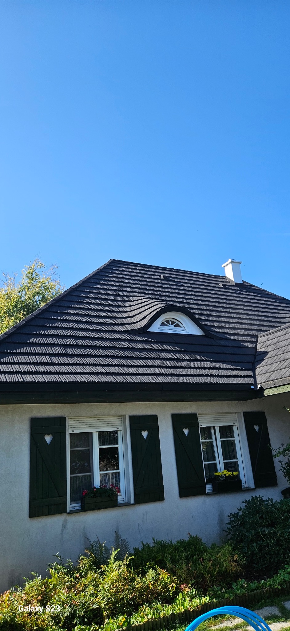 Czarny dach z oryginalnym oknem w kształcie oka na tle błękitnego nieba. Okiennice z serduszkami. Detal architektury domu jednorodzinnego.