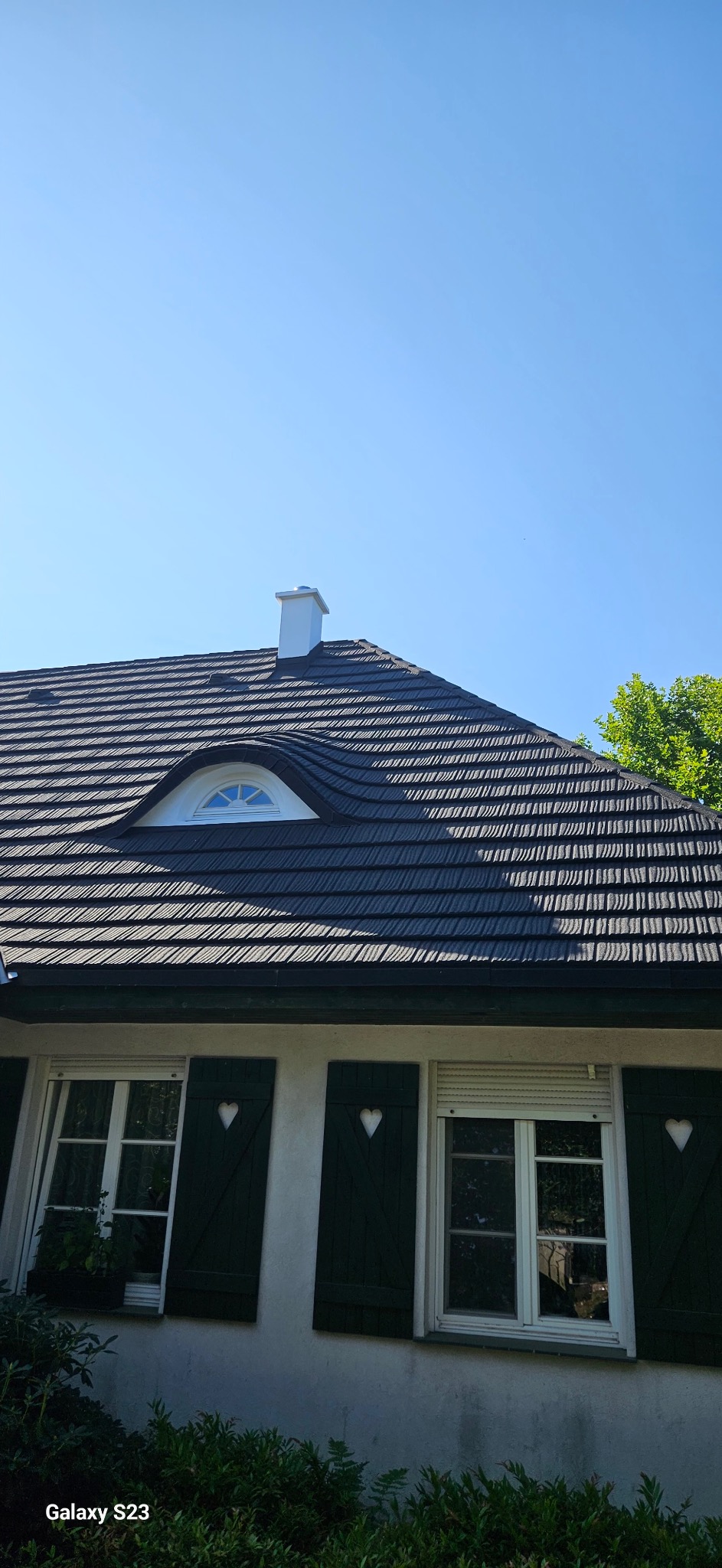 Ciemny dach z dachówki bitumicznej z oknem w kształcie półksiężyca i białym kominem na tle błękitnego nieba. Okna z zielonymi okiennicami z wyciętymi sercami.