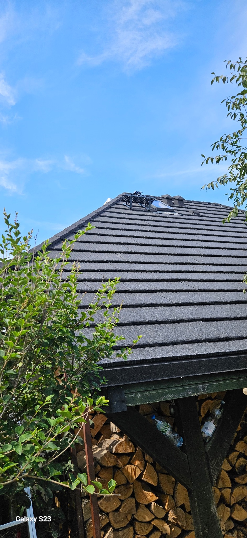Ciemnoszary dach z dachówki modułowej, widoczny fragment okna dachowego i zabezpieczenia przeciwśniegowe. W tle błękitne niebo i zieleń drzew.