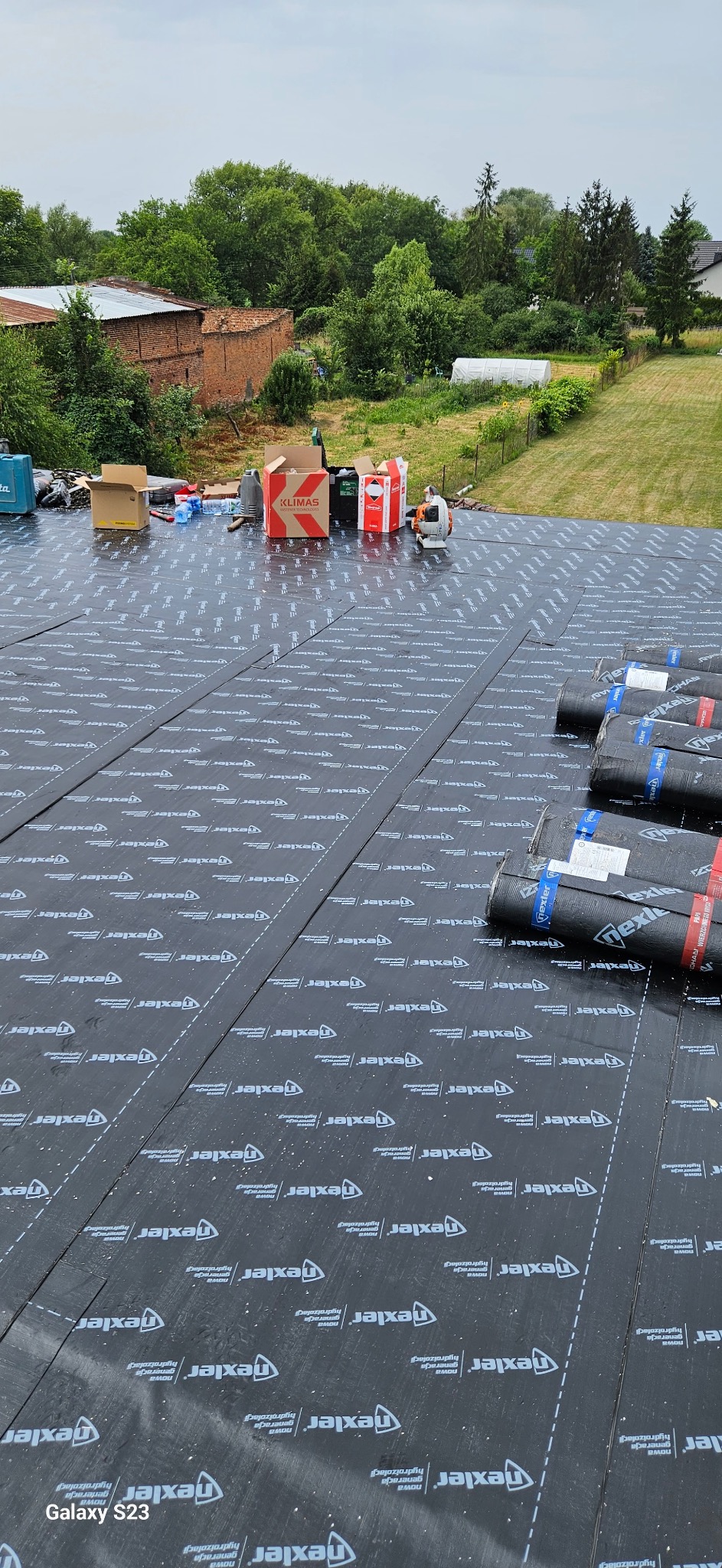 Montaż membrany dachowej Texler na dachu. Widoczne rolki papy, narzędzia i materiały budowlane. W tle zieleń drzew i zabudowania.