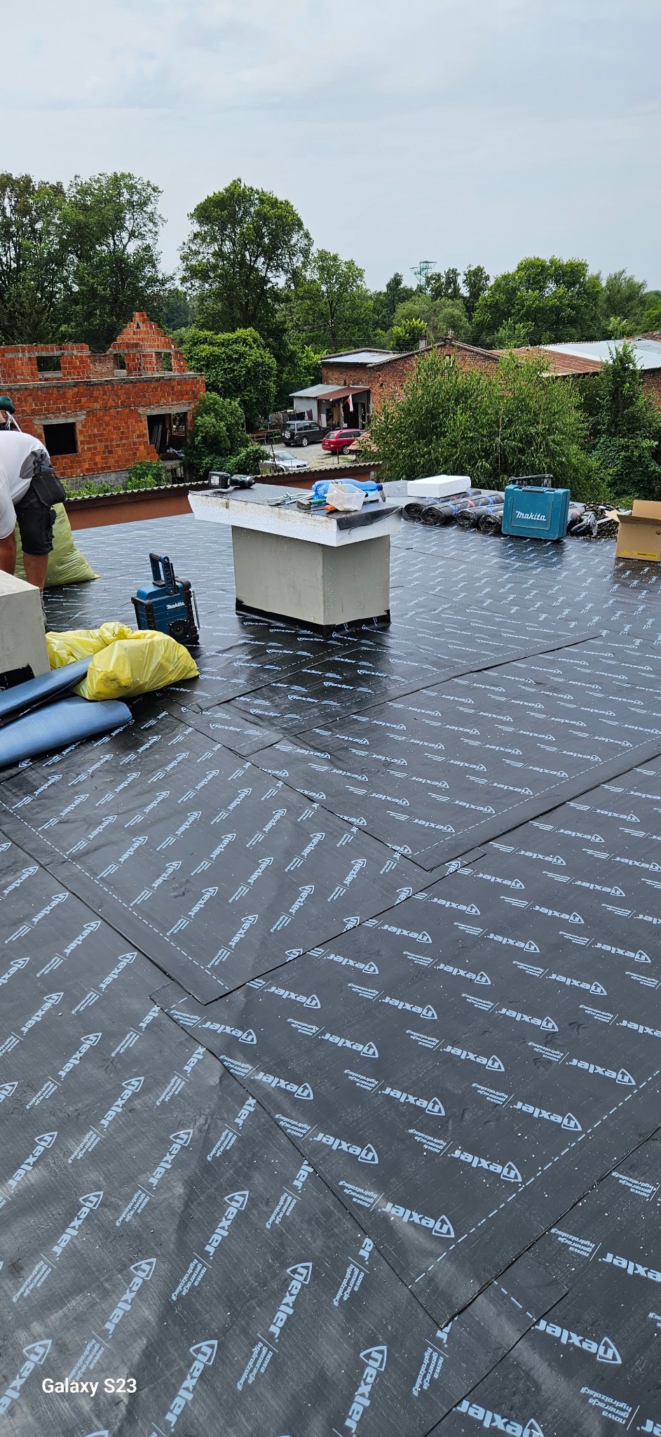 Układanie czarnej membrany dachowej z białym nadrukiem na dachu. Widoczne narzędzia, skrzynka Makita i częściowo widoczny dekarz. W tle ceglany budynek w budowie.