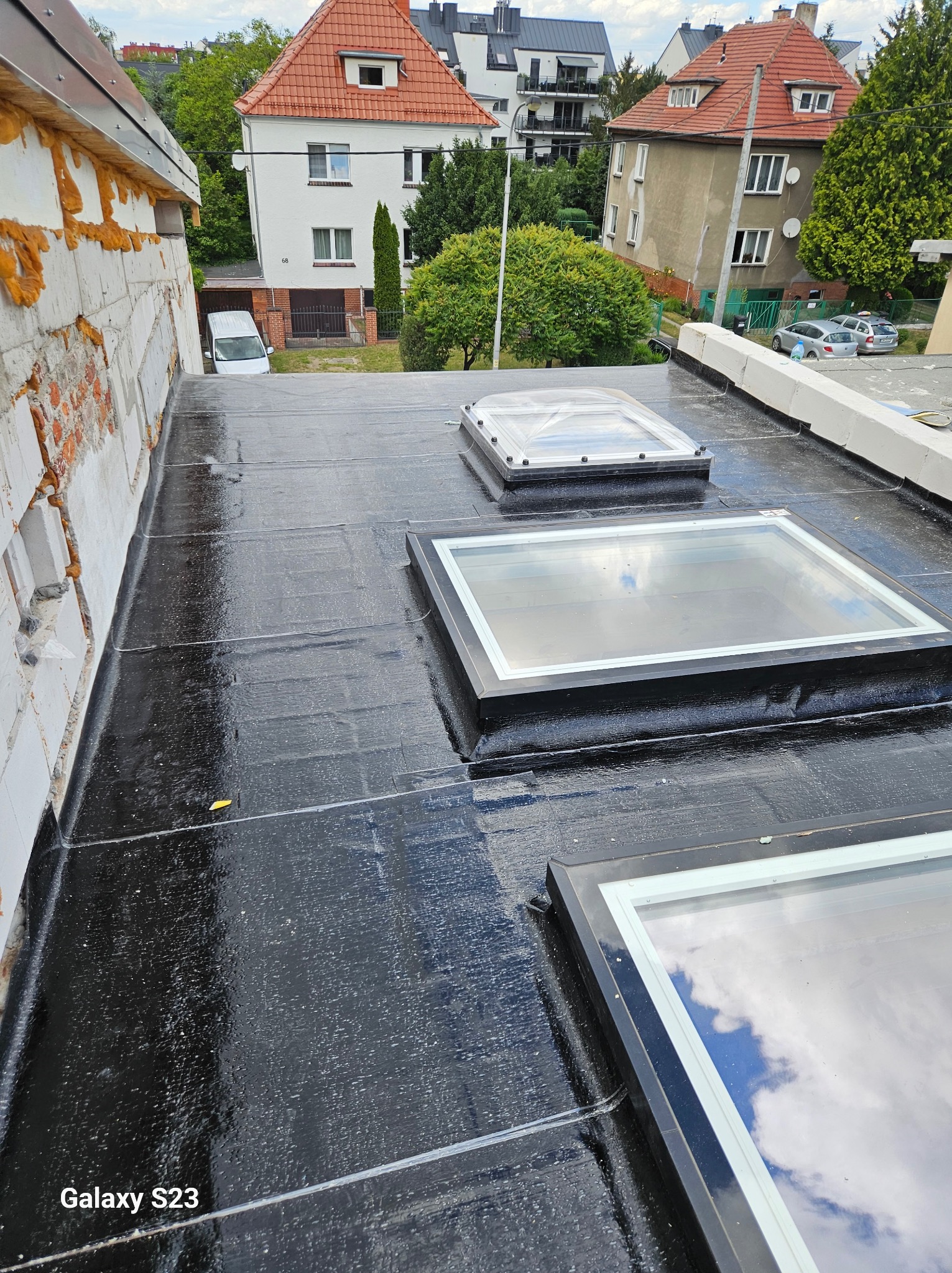 Wykończony dach pokryty czarną papą z trzema oknami dachowymi. Widoczne budynki w tle i fragment ściany z ociepleniem.
