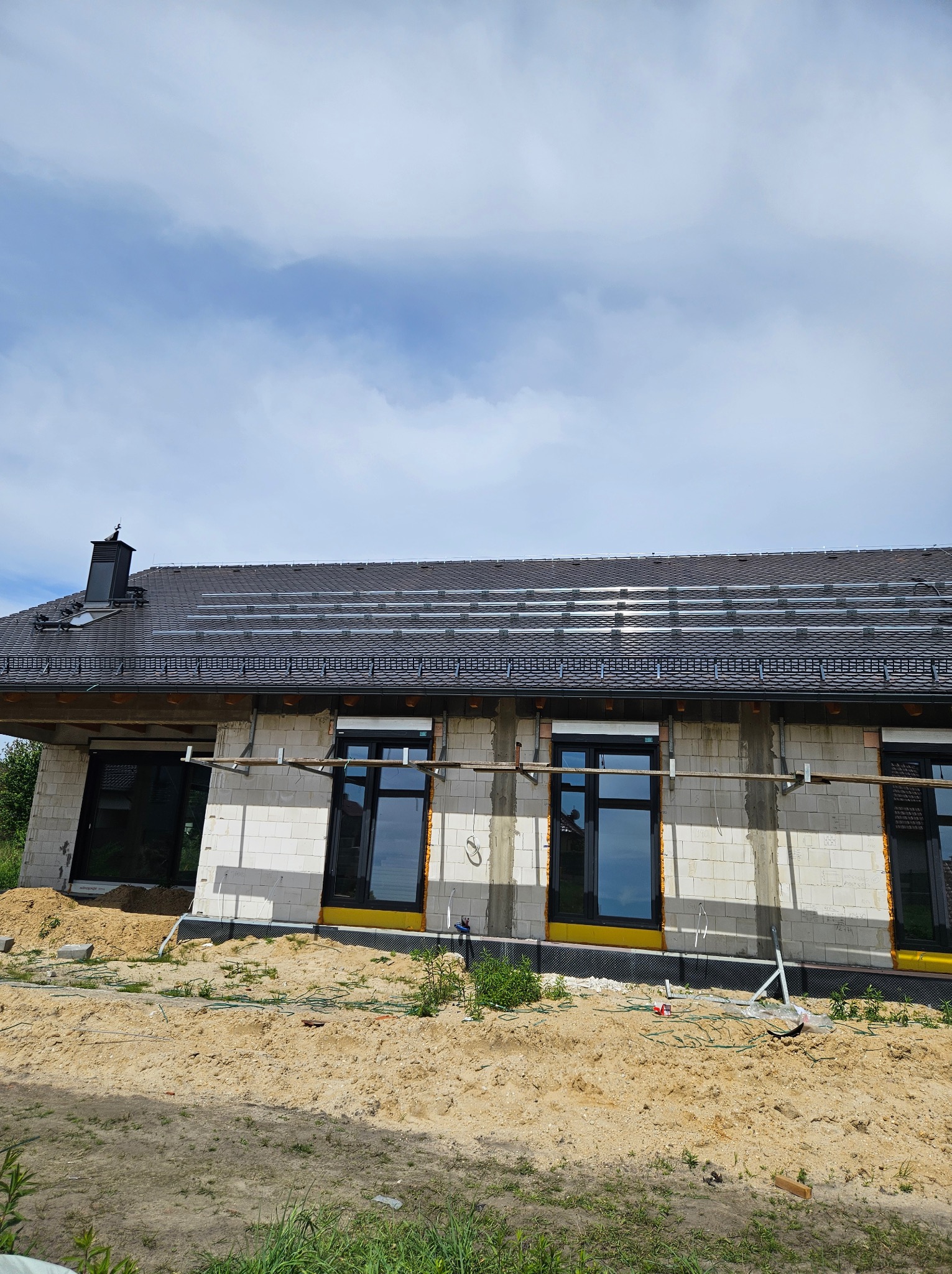 Montaż paneli fotowoltaicznych na dachu domu w trakcie budowy. Widoczne rusztowania i elementy konstrukcyjne. Dachówka w kolorze grafitowym.