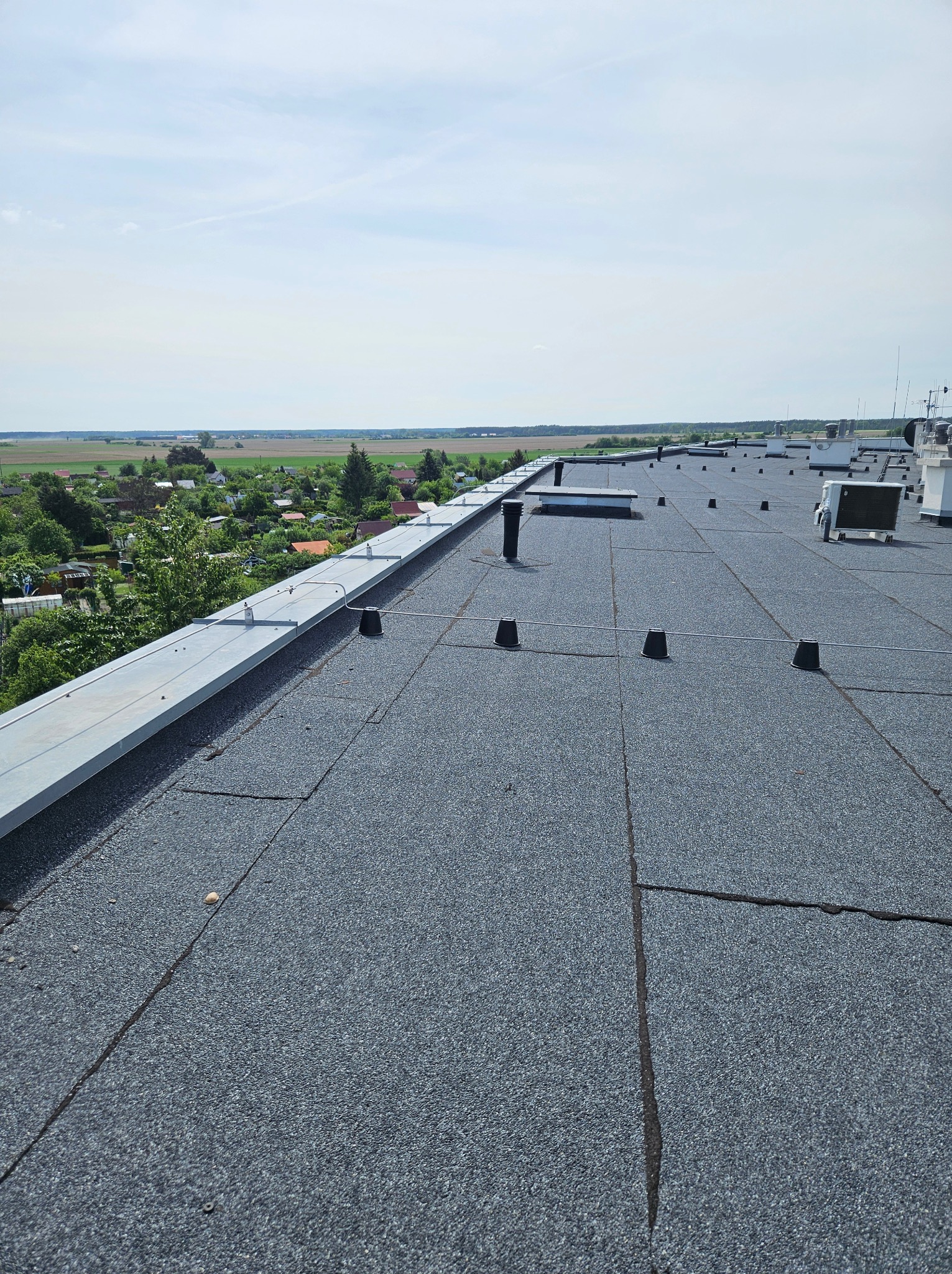 Widok z góry na płaski dach pokryty papą z elementami wentylacyjnymi i zabezpieczeniami, w tle zielony krajobraz i błękitne niebo.