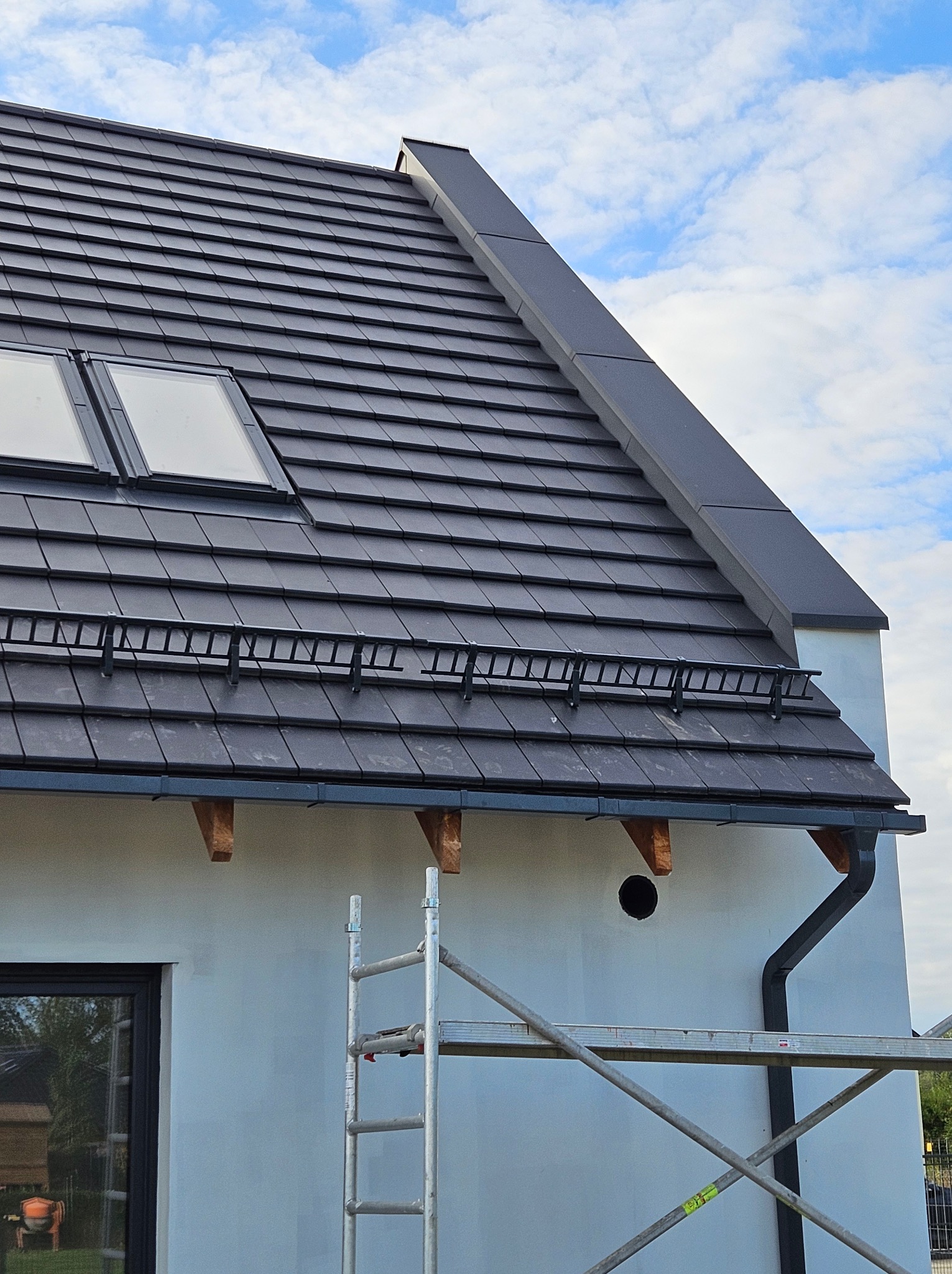 Szary dach z dachówką ceramiczną, okna dachowe, zabezpieczenia przeciwśniegowe i rynna. Fragment ściany budynku z oknem i rusztowaniem w tle.