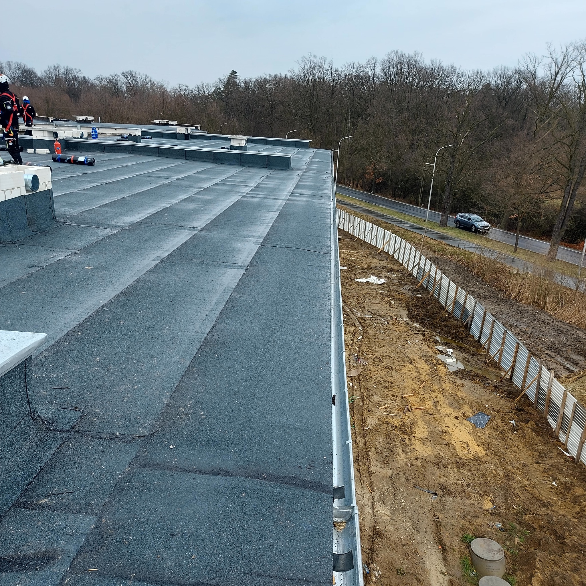 Wykończony, płaski dach pokryty papą termozgrzewalną z widocznymi łączeniami. W tle drzewa i droga z przejeżdżającym samochodem. Widoczne zabezpieczenia na krawędzi dachu.