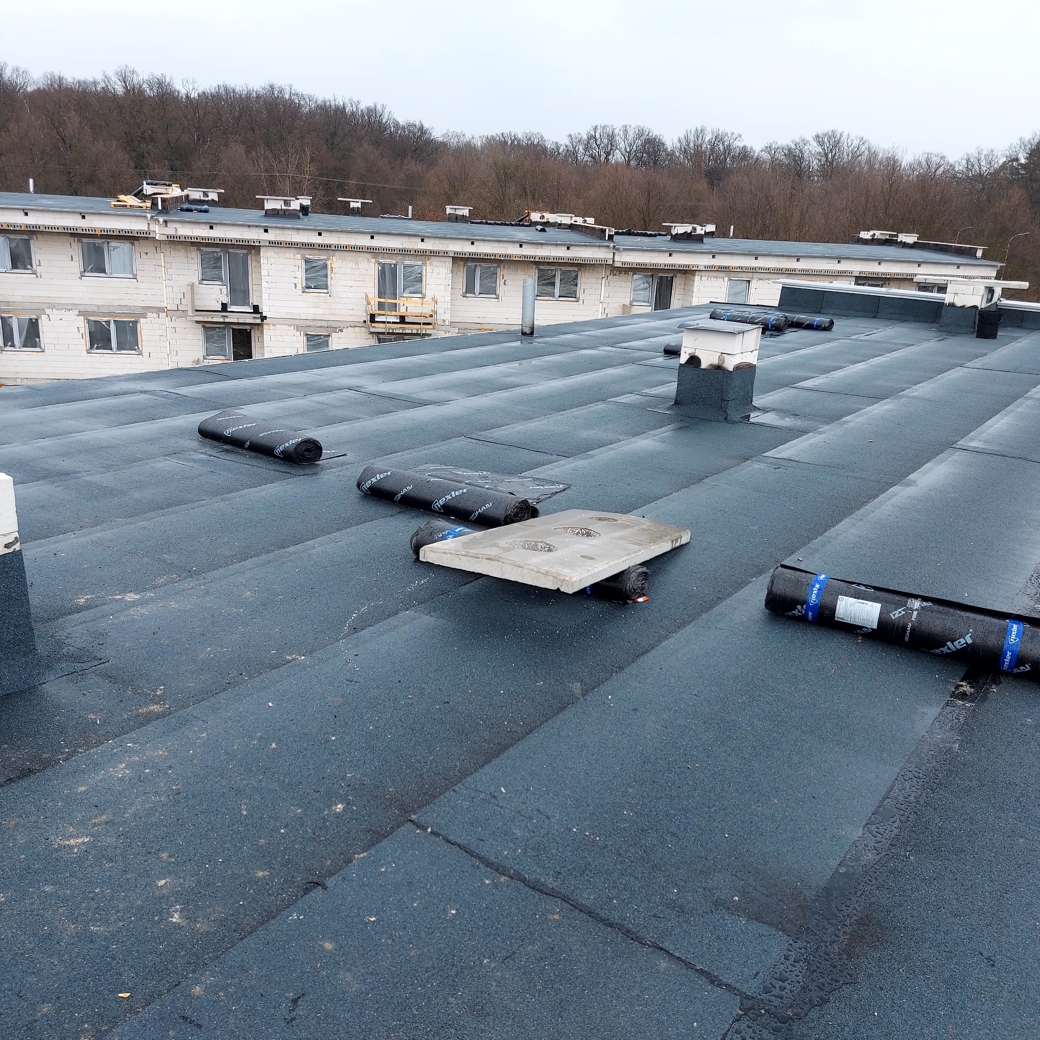 Wykończony płaski dach pokryty papą termozgrzewalną z widocznymi rolkami papy i elementami wentylacyjnymi. W tle budynek w trakcie budowy i drzewa.