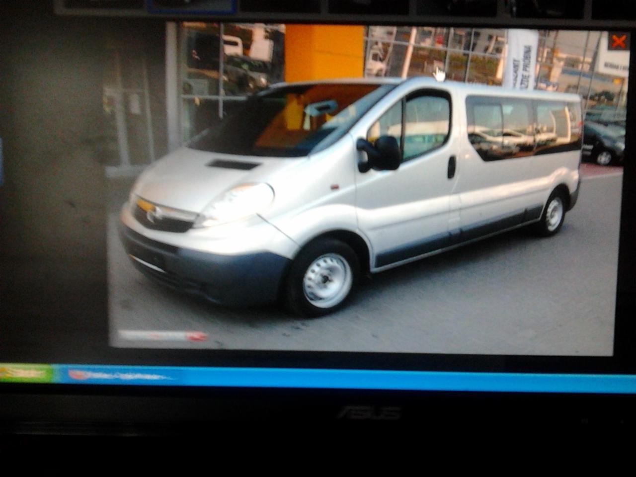 Srebrny Opel Vivaro z ciemnoszarym dołem, zaparkowany przed budynkiem z witrynami i żółtym elementem elewacji.