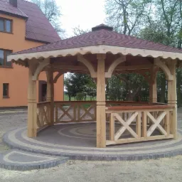 Drewniana altana z czerwonym dachem i ozdobnymi balustradami na okrągłej, wybrukowanej podstawie. W tle budynek mieszkalny i zieleń.