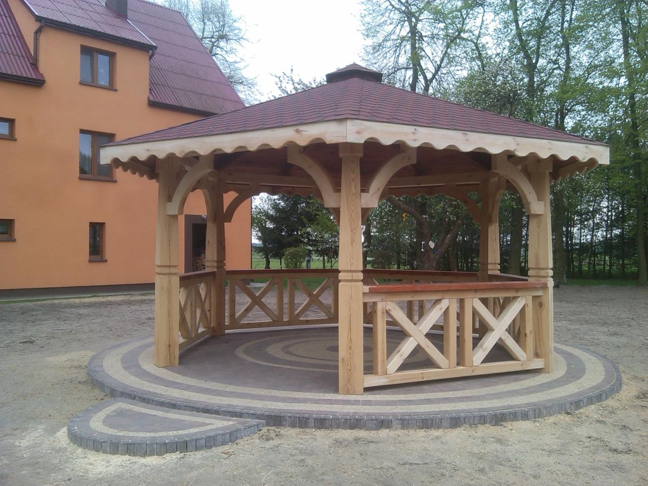 Drewniana altana z czerwonym dachem i ozdobnymi balustradami na okrągłej, wybrukowanej podstawie. W tle budynek mieszkalny i zieleń.