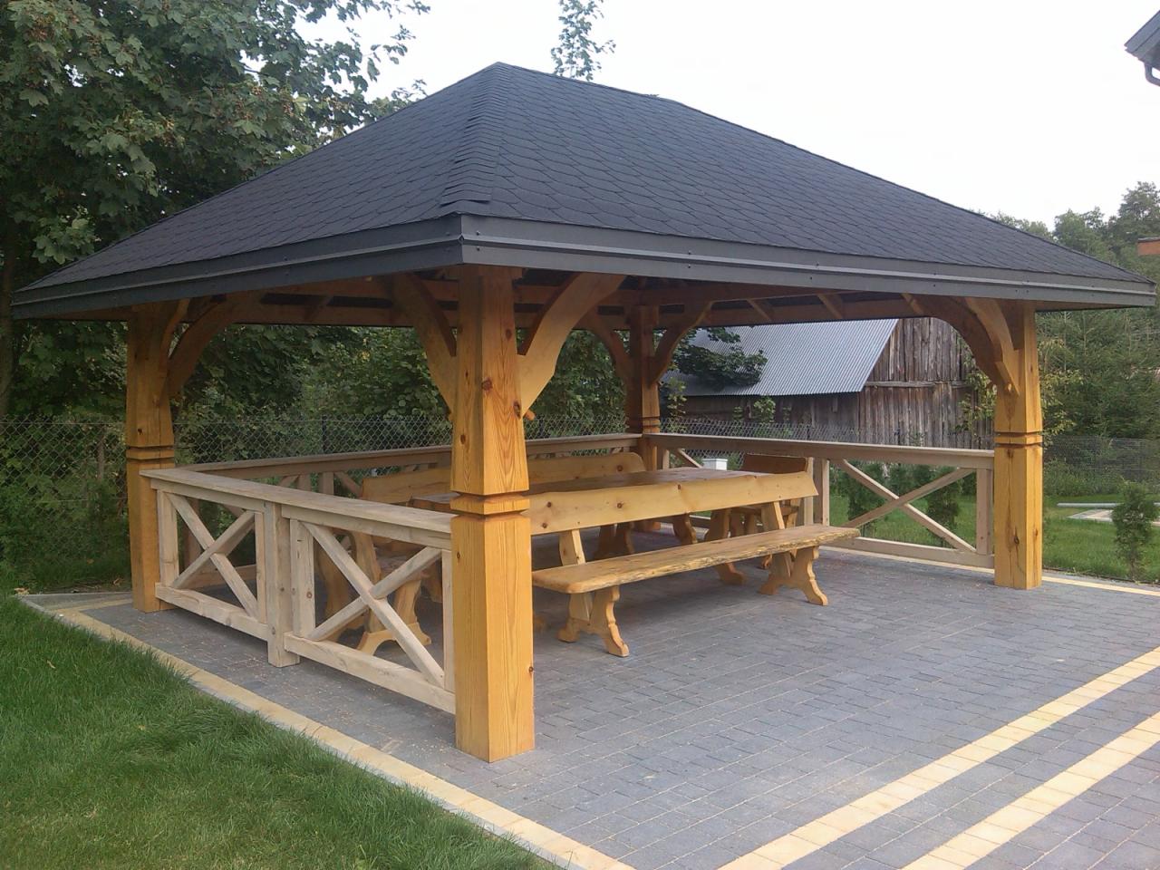 Drewniana altana z czarnym dachem i stołem z ławkami, otoczona kostką brukową. Widoczne detale konstrukcyjne i ogrodzenie z krzyżowych elementów. Tło: budynek gospodarczy.